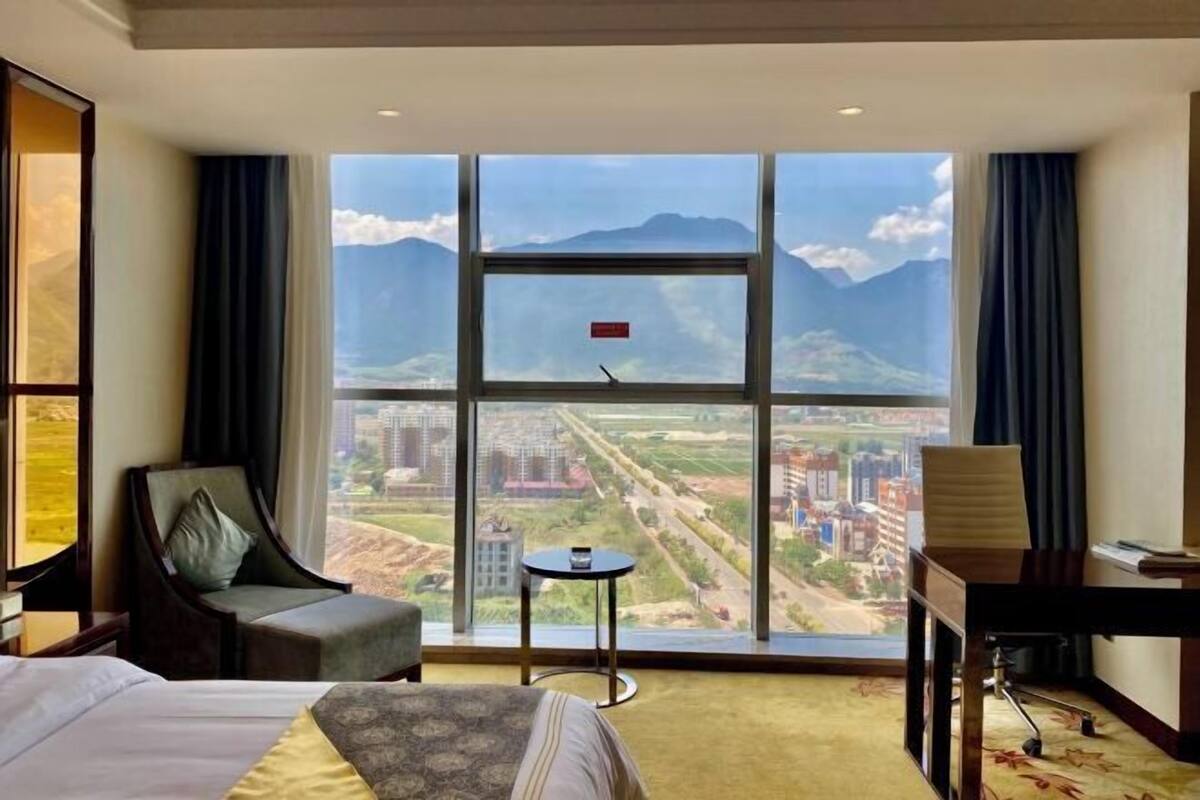 A hotel room with a large window offering a mountain view, a bed, a chair, a small table, and a desk.