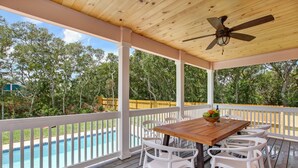 Une terrasse extérieure couverte, avec une table en bois et des chaises blanches, donnant sur une piscine et des arbres.