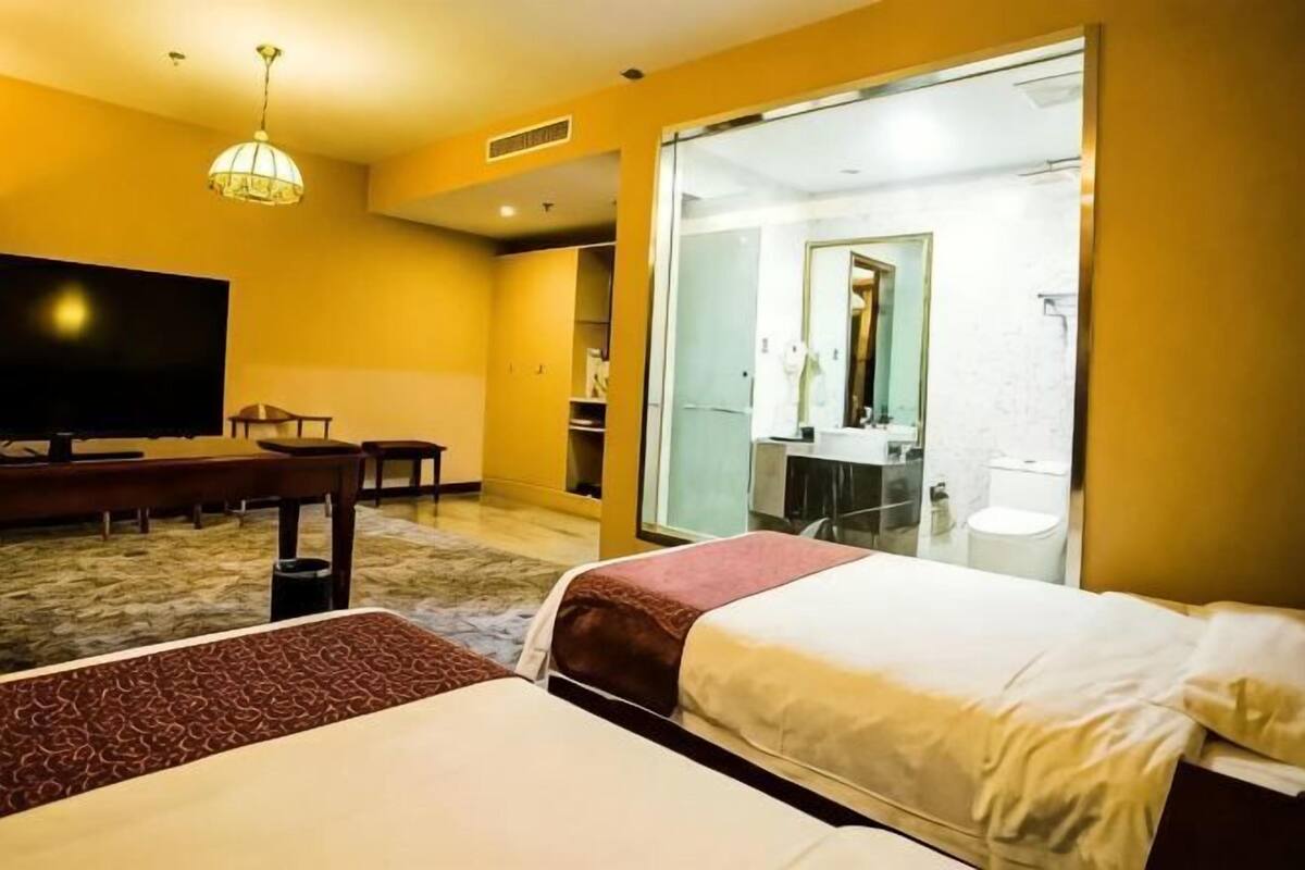 A hotel room with a bed, a desk, a television, and a bathroom visible through a glass door.