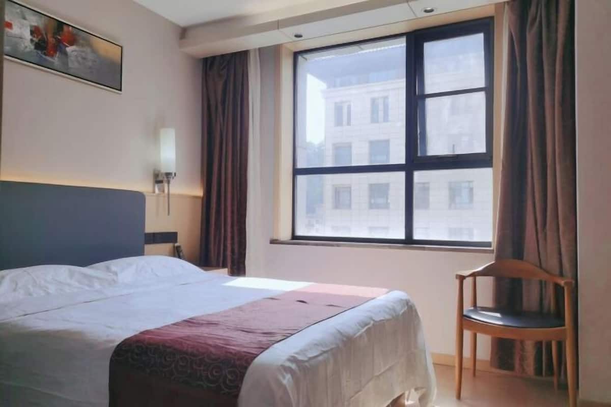 A hotel room with a bed, a chair, a window with curtains, and a wall-mounted lamp.
