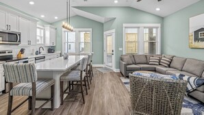 A modern kitchen with white cabinets, a central island with chairs, a ceiling fan, and a dining area with a sofa and a wicker basket. - Brand New Single Level Condo, Steps from the Beach  (Corolla)