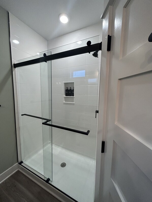 A modern bathroom with a glass shower enclosure, a black towel rack, and a small shelf with toiletries. - Brand New Single Level Condo, Steps from the Beach (Corolla)