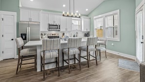 A kitchen with white cabinets, a stainless steel refrigerator, a microwave, and a dining area with striped chairs. - Brand New Single Level Condo, Steps from the Beach  (Corolla)