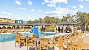A poolside area with wooden lounge chairs and tables, a covered seating area, and a pool. - Brand New Single Level Condo, Steps from the Beach  (Corolla)
