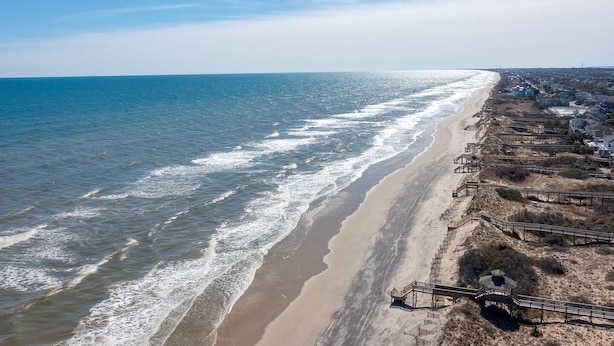 A coastal beach with a wooden boardwalk leading to a sandy area, and buildings along the shore. - Brand New Single Level Condo, Steps from the Beach  (Corolla)
