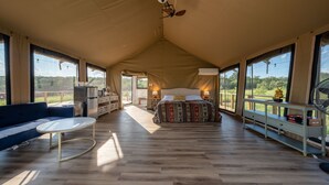 A spacious tented room with a bed, a sofa, a table, and large windows offering a view of the outdoors.