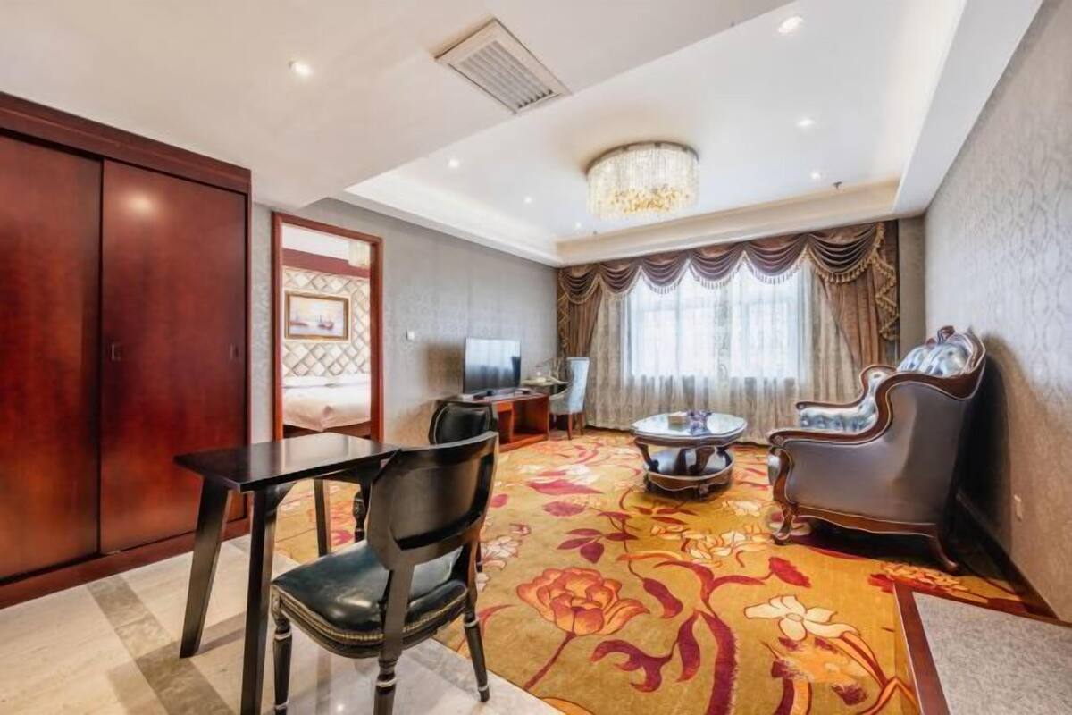 Een hotelkamer met een eettafel, stoelen, een kroonluchter, een televisie en een groot raam met gordijnen.