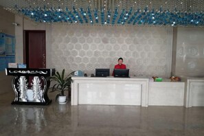 Een receptie met een baliemedewerker, een receptiebalie, een decoratieve plant en een behang met patroon.