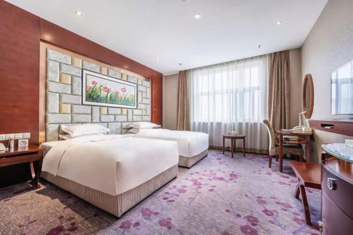 Een hotelkamer met twee bedden, een klein bureau, een stoel, een spiegel en een raam met gordijnen.