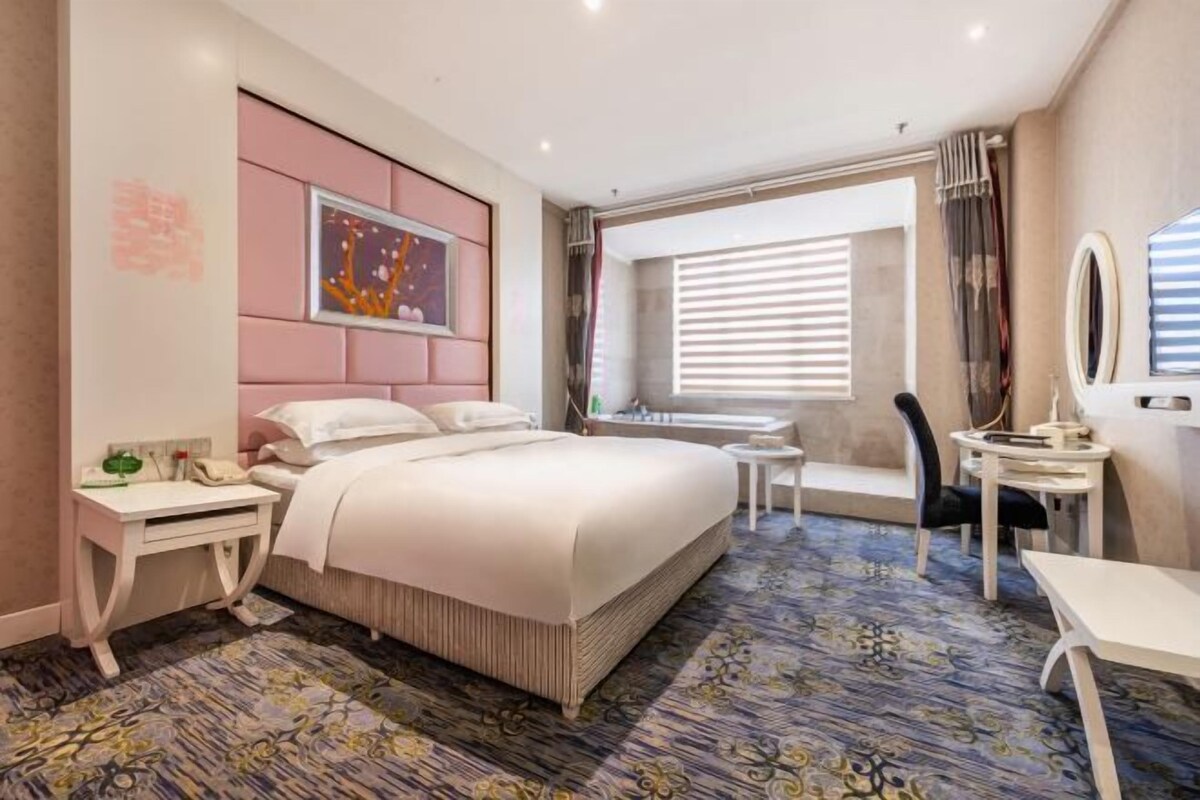 Une chambre d’hôtel comprenant un lit, des tables de chevet, un bureau, une chaise, une fenêtre avec des stores et un tableau au mur.
