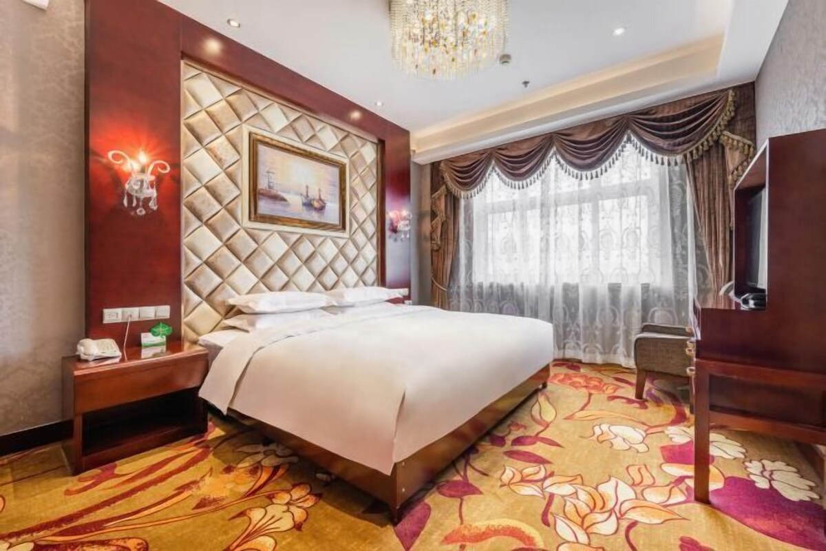 Une chambre d’hôtel avec un grand lit, une table de chevet, un bureau, un lustre et une fenêtre avec des rideaux.