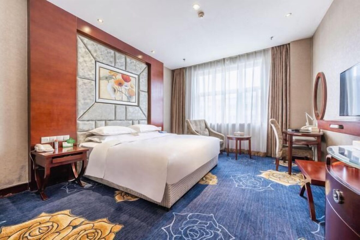 Une chambre d’hôtel avec un grand lit, un bureau, une chaise, une télévision et une fenêtre avec des rideaux.