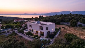Un edificio moderno blanco con techo plano, rodeado de vegetación y vistas a un lago al atardecer.