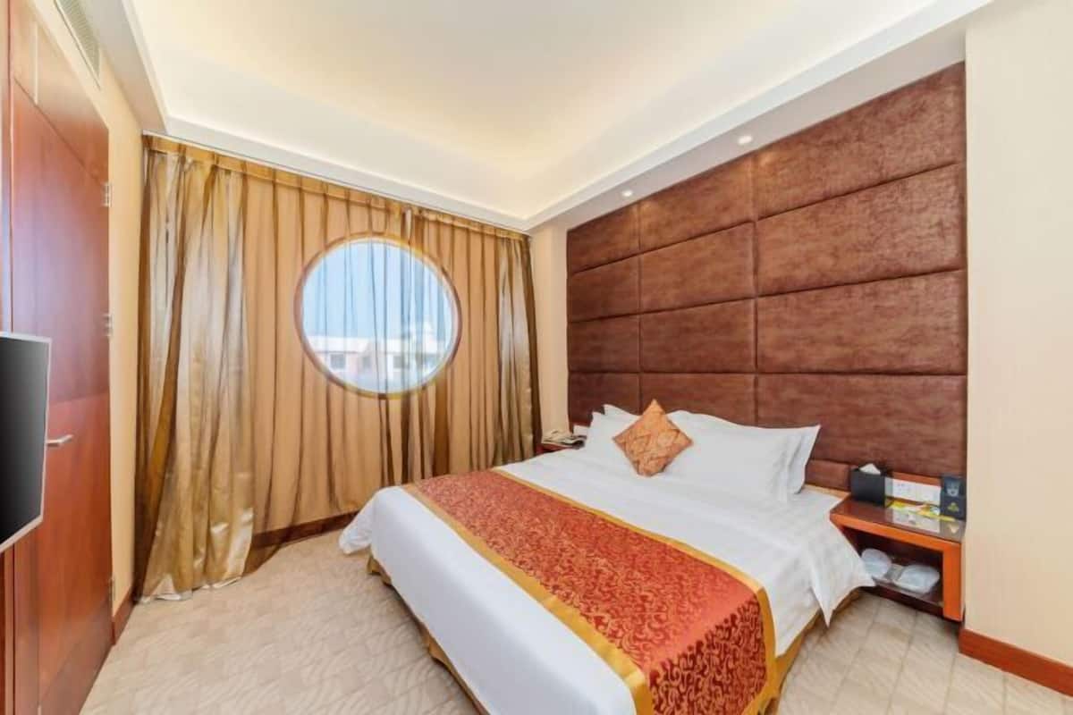 Une chambre d’hôtel avec un grand lit, une fenêtre ronde, une tête de lit en bois et un téléviseur placé à gauche.