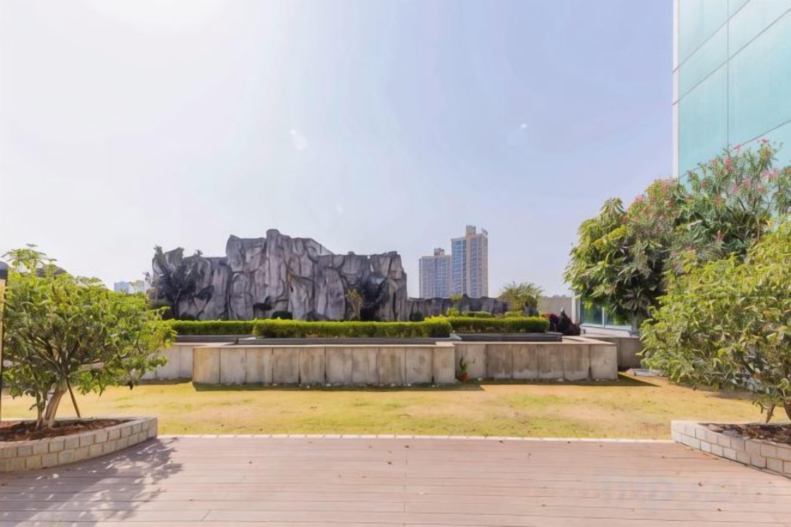Un área ajardinada con una formación rocosa, un sendero pavimentado y un edificio al fondo.