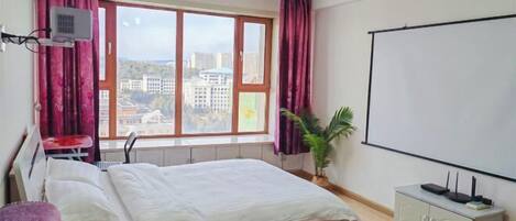 Una habitación de hotel con cama, escritorio, ventana con cortinas, una planta y una pantalla para proyector.