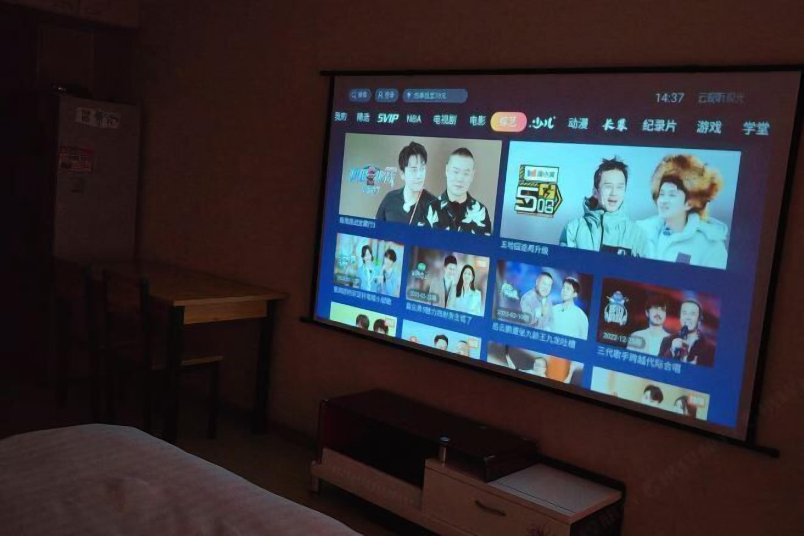A hotel room with a large TV screen displaying a variety of content, a wooden table, and a small wooden cabinet.