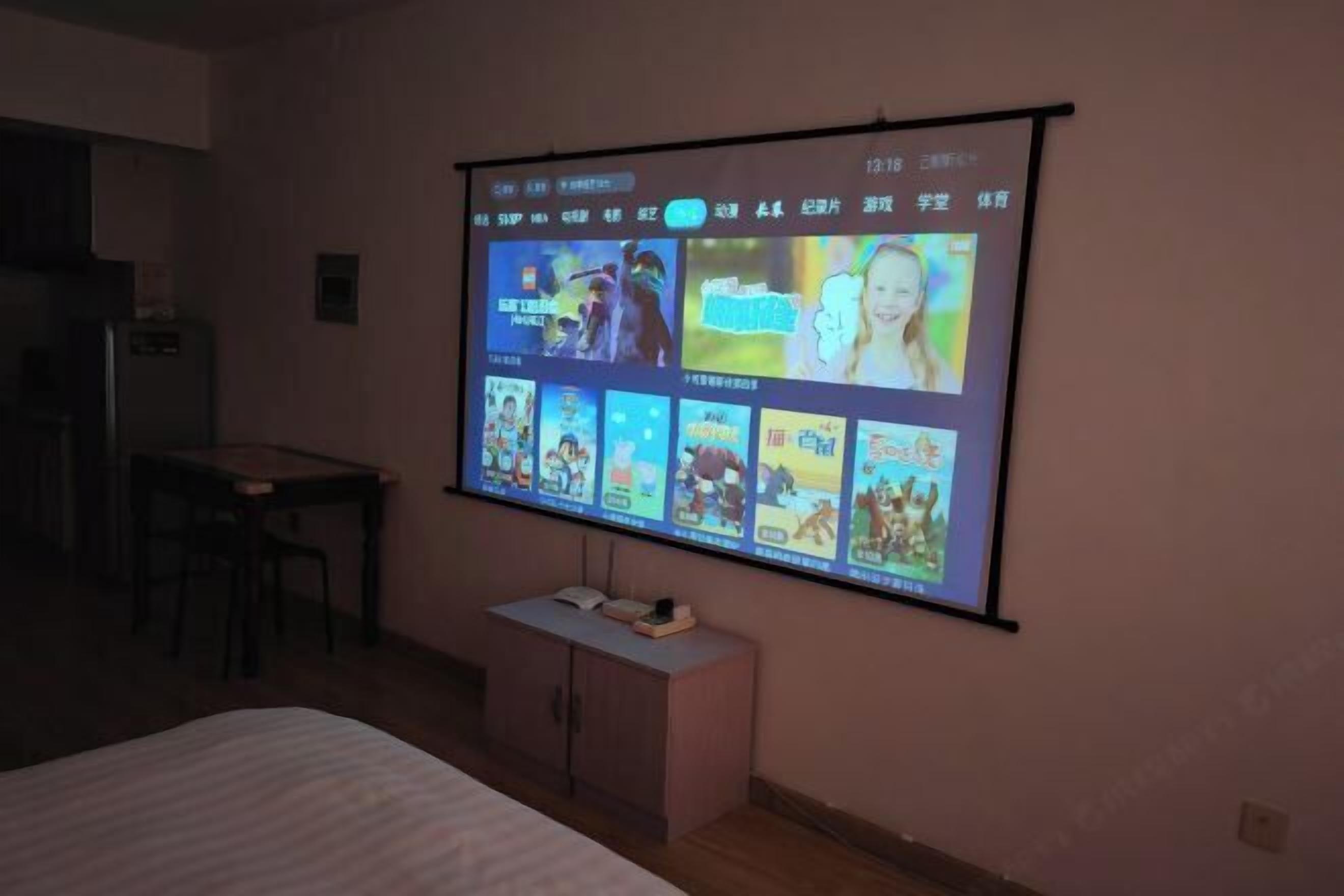 A room with a bed, a desk, a chair, and a wall-mounted projector screen displaying various images.