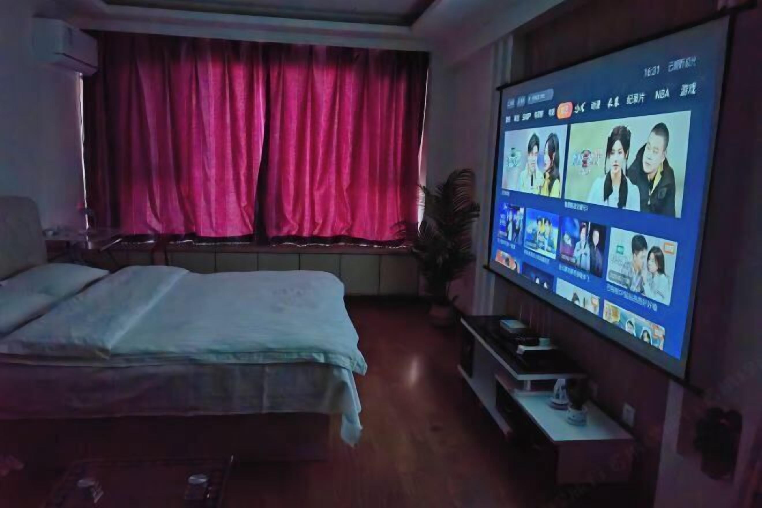 A bedroom with a bed, a TV mounted on the wall displaying a video call interface, a window with curtains, and a small table with a plant.