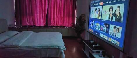 A bedroom with a bed, a TV mounted on the wall displaying a video call interface, a window with curtains, and a small table with a plant.