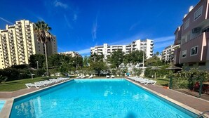 Una piscina rectangular con borde de baldosas, rodeada de sillas de descanso y sombrillas, con edificios altos al fondo.