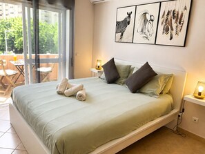 A bed with a light green bedspread, two pillows, and rolled towels. There are wall art pieces above the bed and a small table with a lamp beside it. - Charming duplex apartment with a terrace and a view of the pool, 200 m from the beach (Portimão)