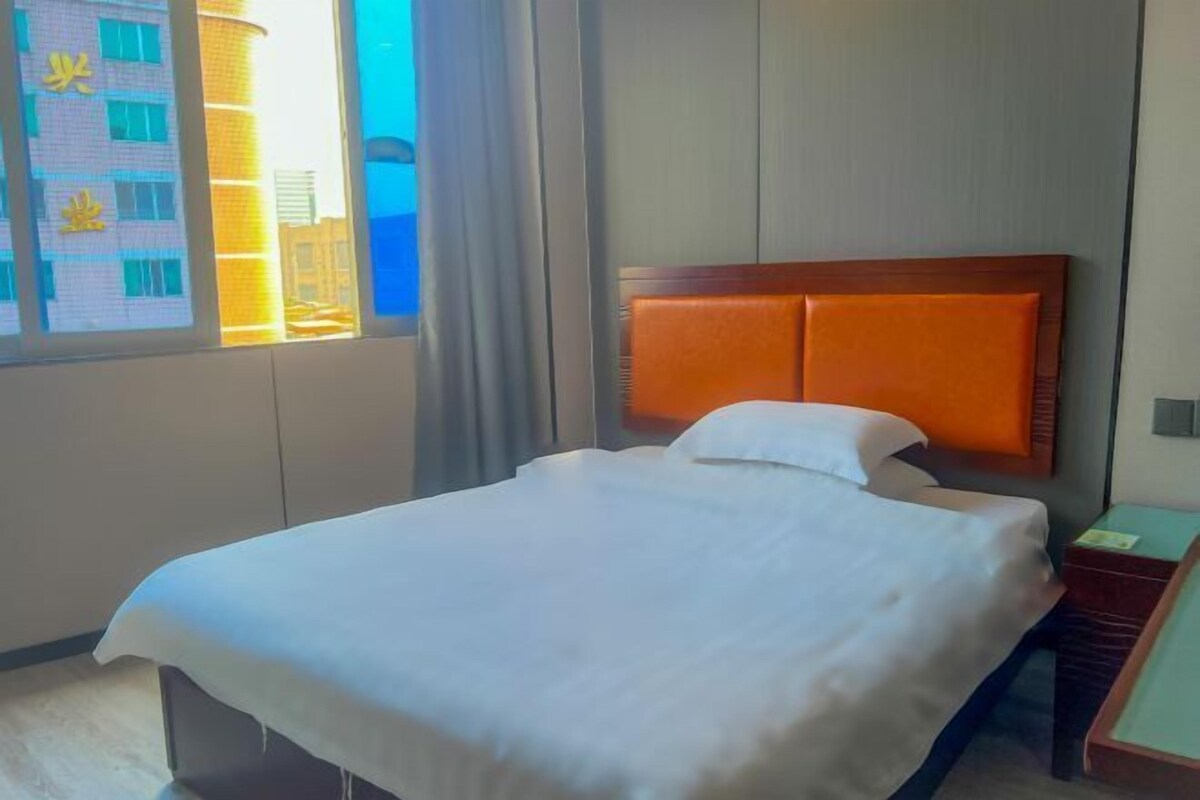 A hotel room with a large bed, a wooden headboard, a view of a building through the window, and a small table with a lamp.