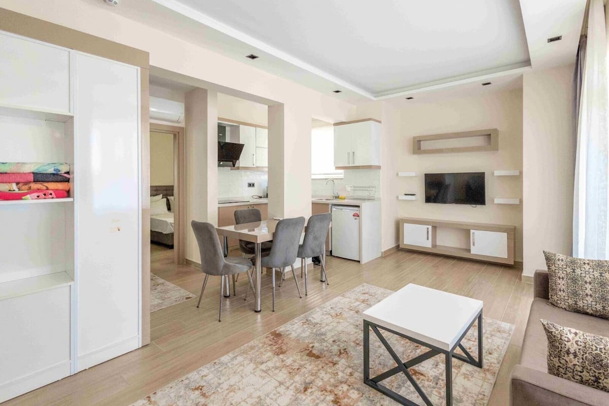 A modern living room with a dining area, a TV mounted on the wall, and a kitchen in the background.