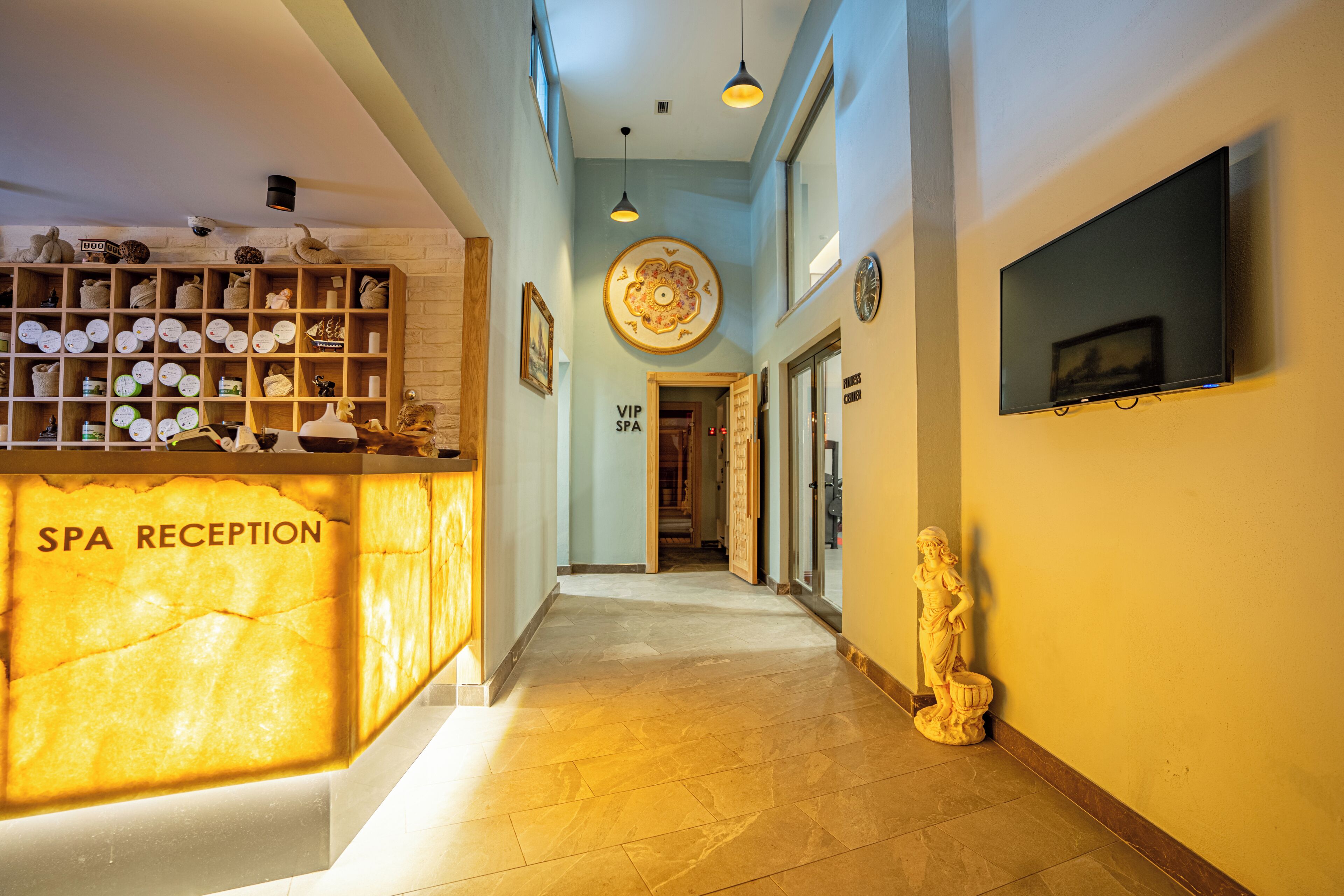 A hotel reception area with a reception desk labeled 'SPA RECEPTION,' a wall-mounted TV, and a statue on the right.
