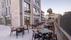Um café ao ar livre com mesas e cadeiras, um edifício com grandes janelas e uma palmeira ao fundo.