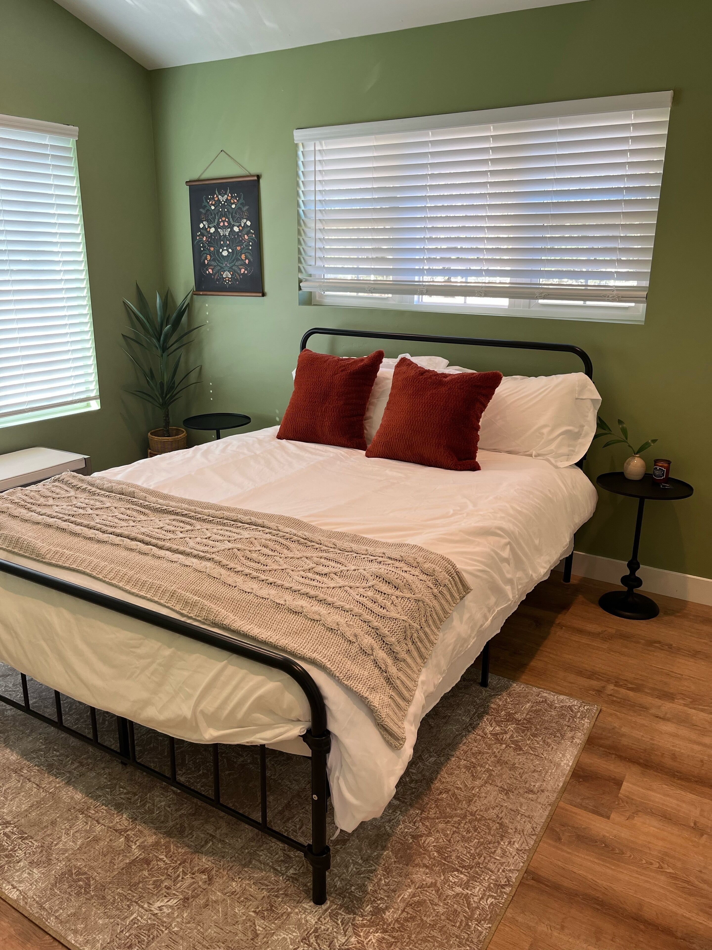 A bedroom with a bed, two pillows, a bedside table, a wall-mounted artwork, and a potted plant.
