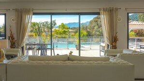 En moderne stue med en stor sofa, et spiseområde med bord og stole, samt udsigt til et pool og bjerge gennem store vinduer.