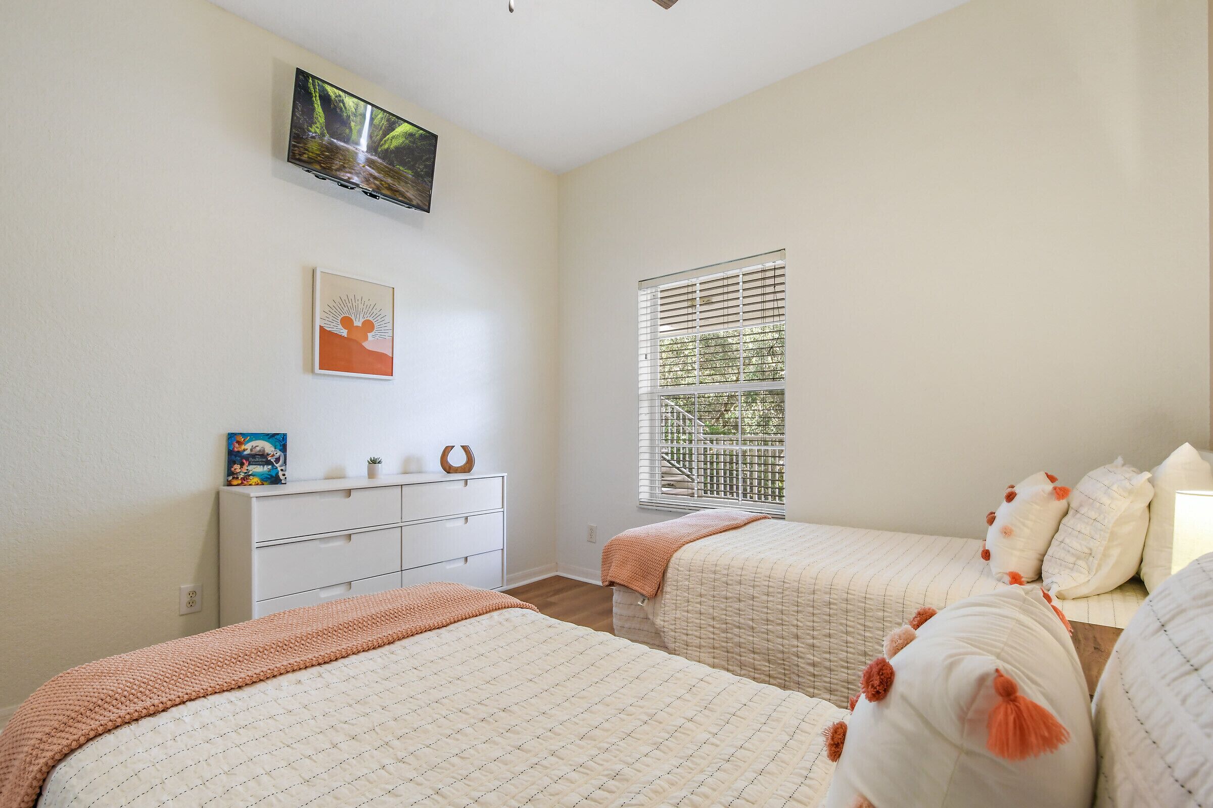 Une chambre avec deux lits, un téléviseur fixé au mur, une fenêtre avec des stores et une commode sur laquelle est posée une petite plante.