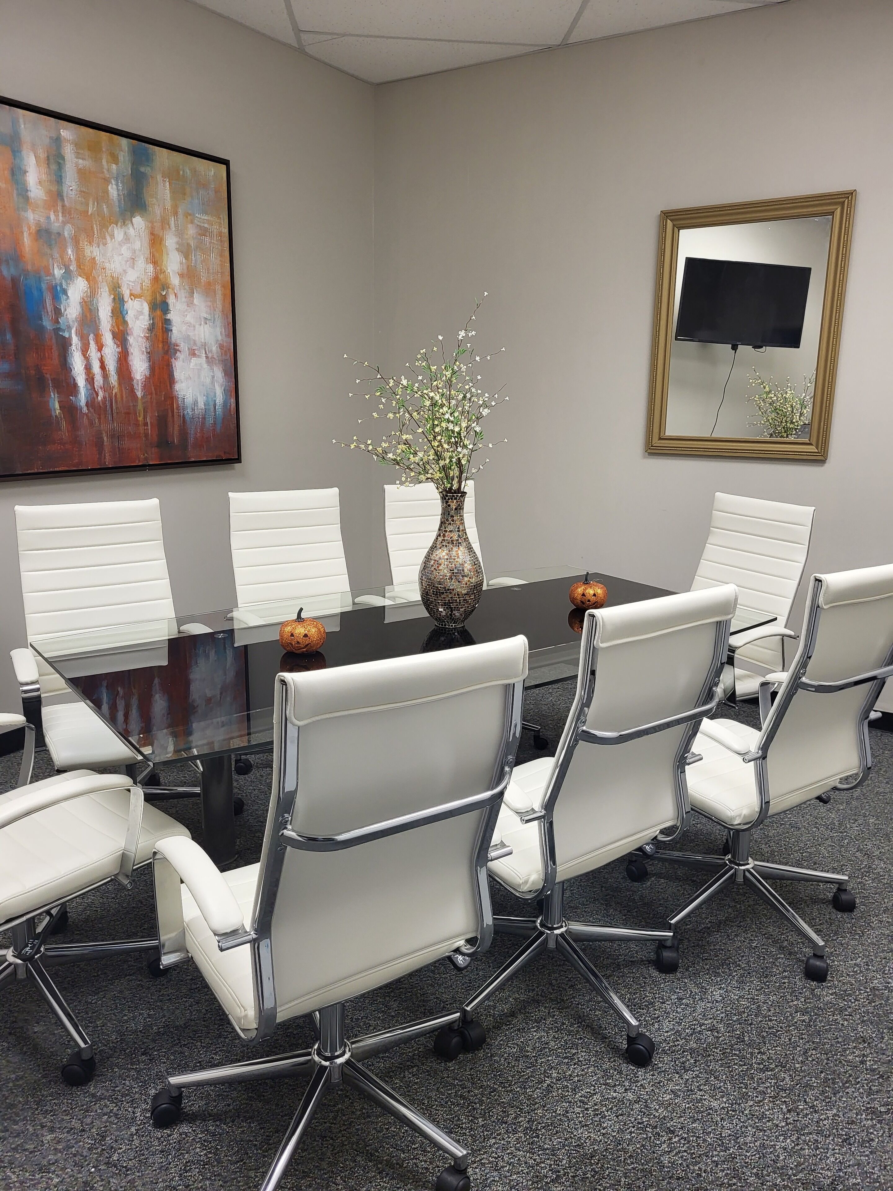A conference room with a glass table, white chairs, a vase with flowers, a painting, and a TV on the wall.