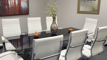 A conference room with a glass table, white chairs, a vase with flowers, a painting, and a TV on the wall.