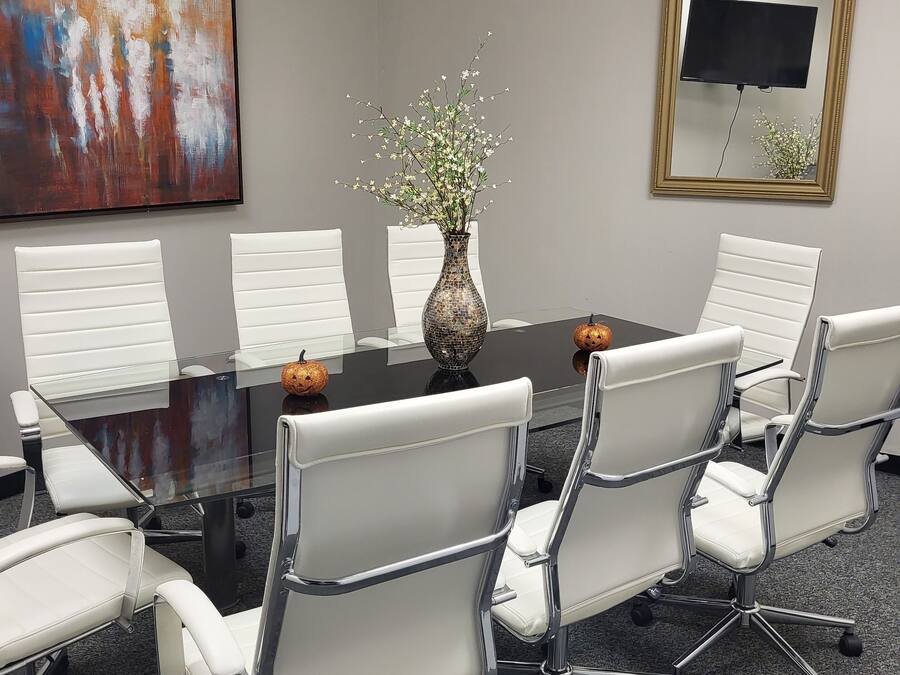 Une salle de conférence avec une table en verre, des chaises blanches, un vase avec des fleurs, un tableau et un téléviseur fixé au mur.