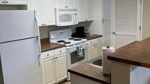 A kitchen with white cabinets, a refrigerator, a microwave, a stove, and a trash can. - Washburn Hotel Suites (Topeka)