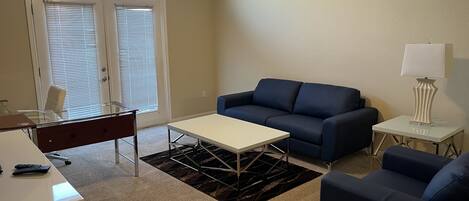 Una sala de estar con un sofá azul, una mesa de centro blanca, un escritorio con una silla y una lámpara sobre una mesita.