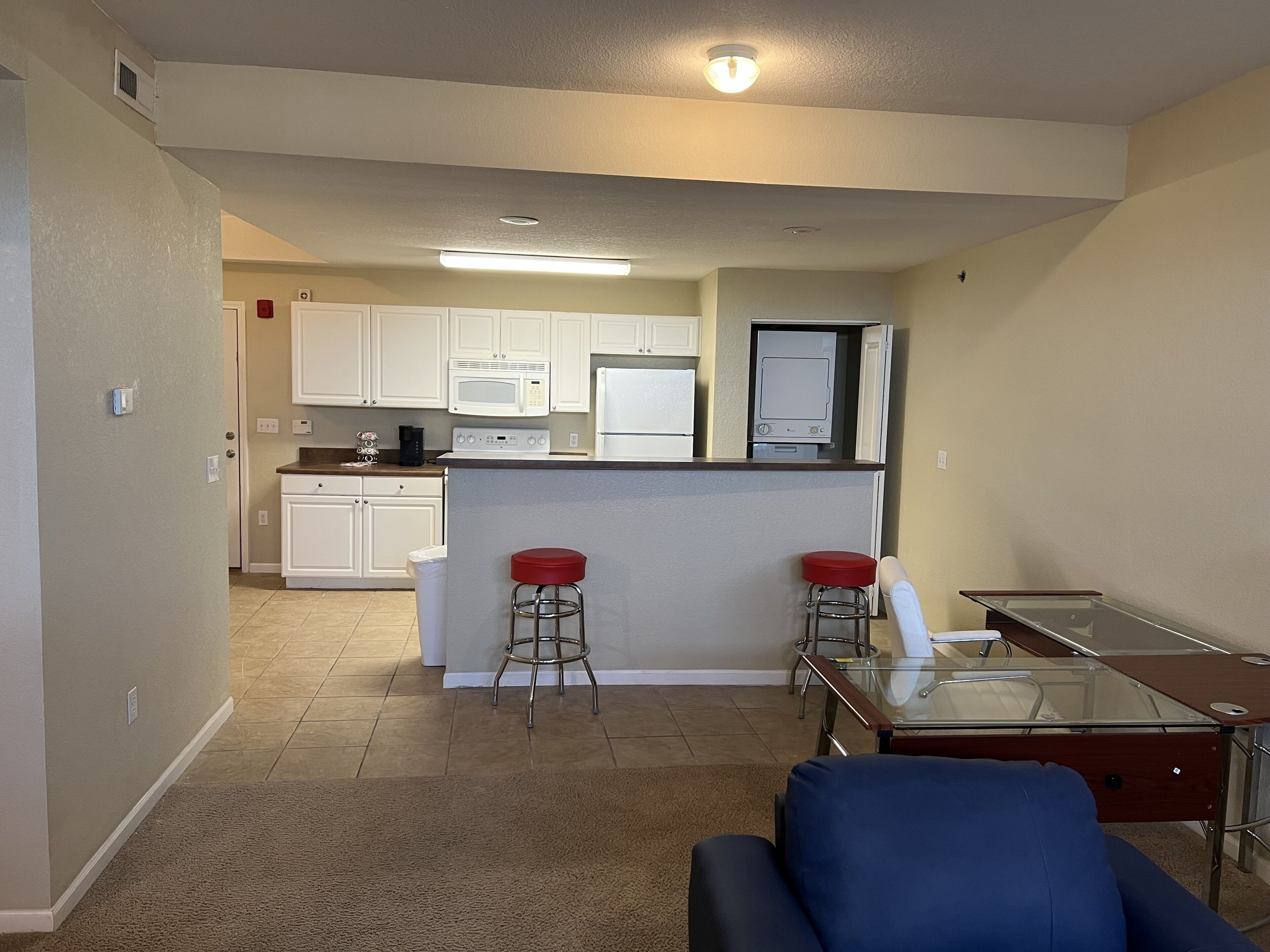 A compact living space with a kitchenette, a glass-top dining table, two red bar stools, and a blue sofa.