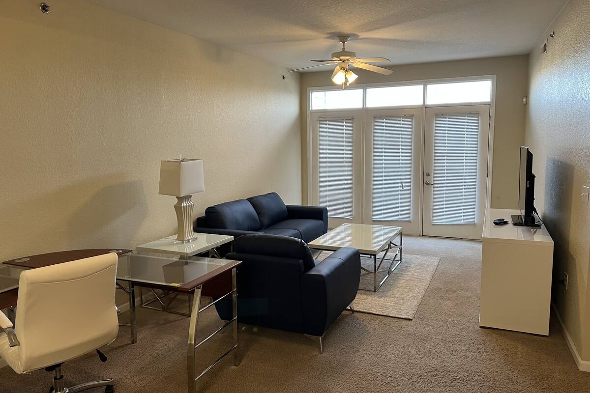 A furnished living room with a sofa, a desk, a chair, a lamp, a ceiling fan, and a television.