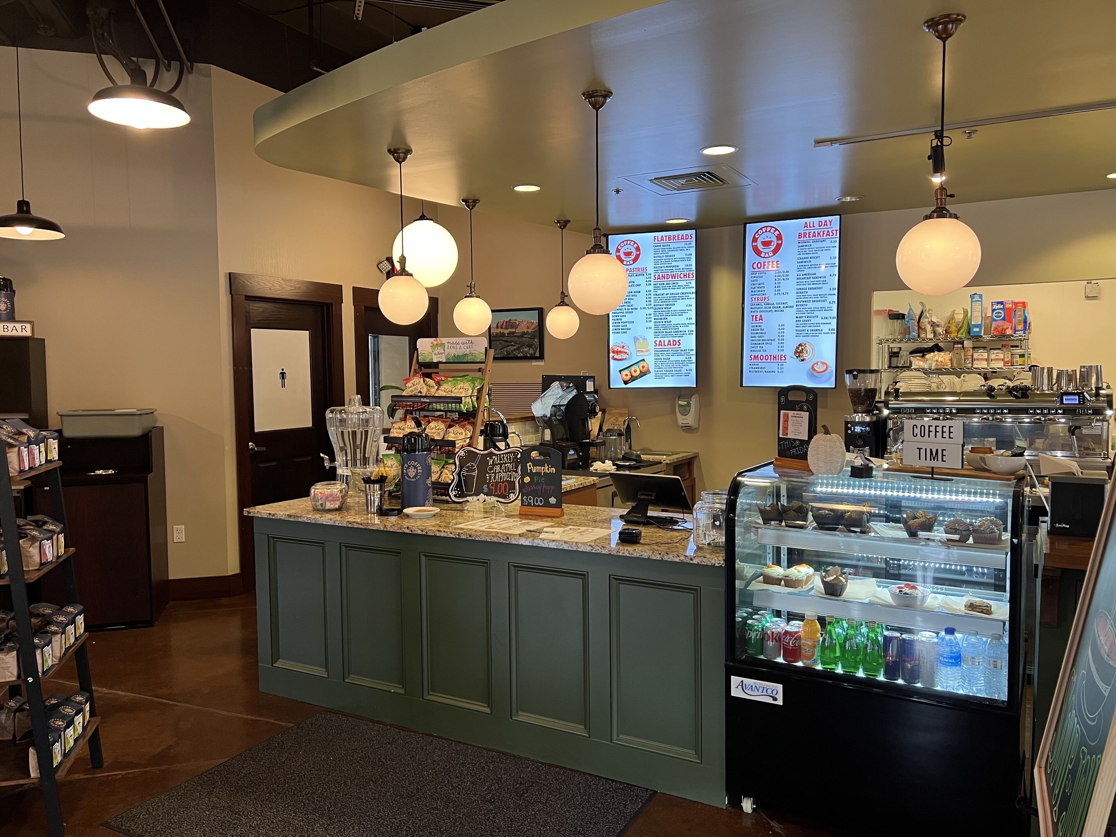 Interior de una cafetería con mostrador, pizarras de menú, dispensador de bebidas y vitrina refrigerada.