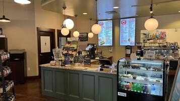 Interior de una cafetería con mostrador, pizarras de menú, dispensador de bebidas y vitrina refrigerada.