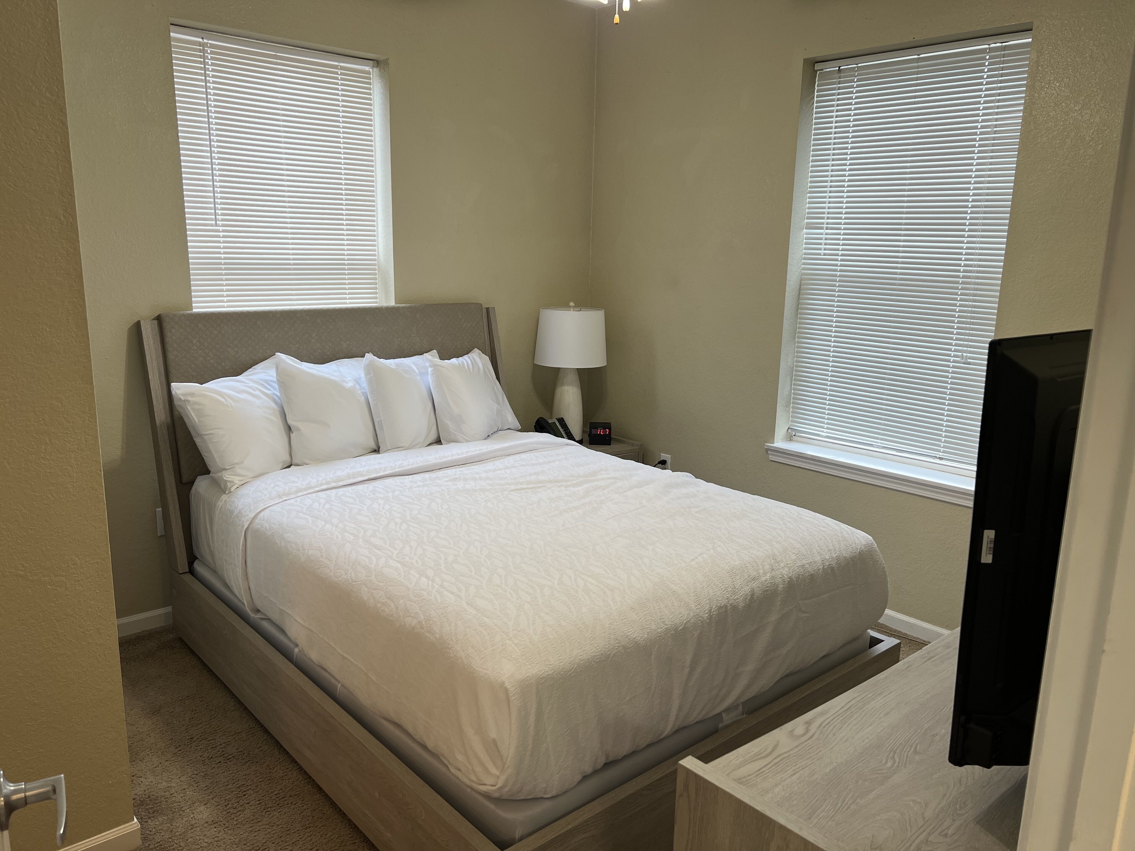 A neatly made bed in a room with two windows, a bedside table with a lamp, and a door to a closet.