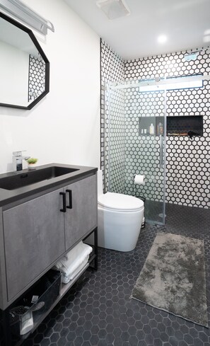 Une salle de bain moderne avec un sol carrelé de motifs hexagonaux noirs et blancs, un meuble-lavabo gris avec un miroir, des toilettes blanches et une cabine de douche en verre.