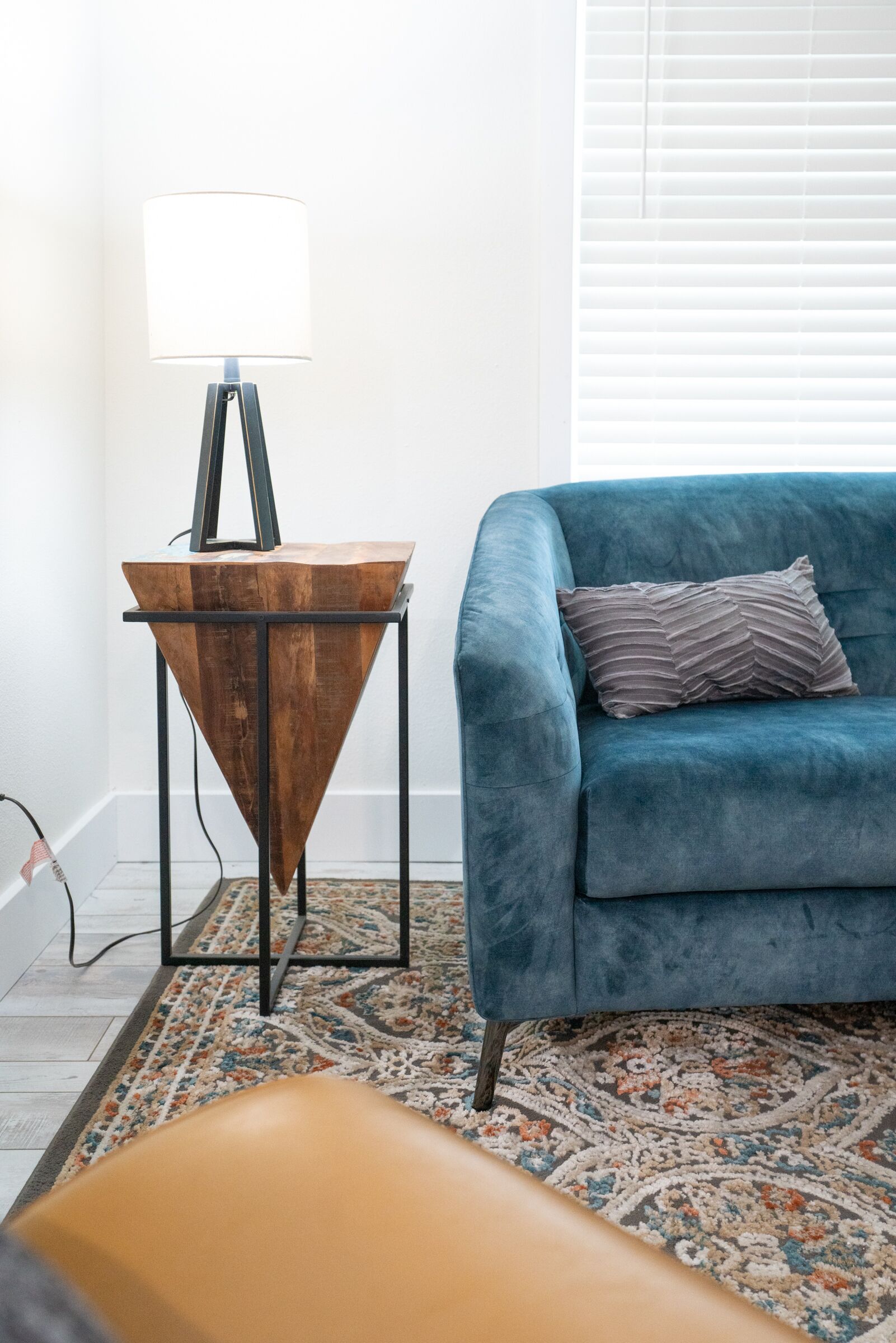 A blue sofa with a patterned pillow, a wooden side table with a lamp, and a patterned rug in a room with white walls and a window with blinds.