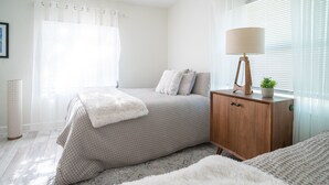 Ein Schlafzimmer mit einem Bett, einem Nachttisch aus Holz mit einer Lampe, einer kleinen Topfpflanze und einem Fenster mit Jalousien.
