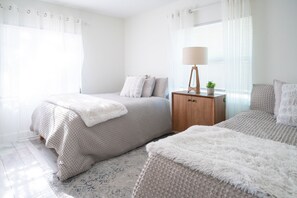 Une chambre avec deux lits, une table de chevet en bois avec une lampe et une fenêtre ornée de rideaux légers.
