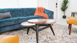 Ein Wohnzimmer mit einem blauen Sofa, zwei runden Holztischen, einem gemusterten Teppich und einem gelben Sessel.