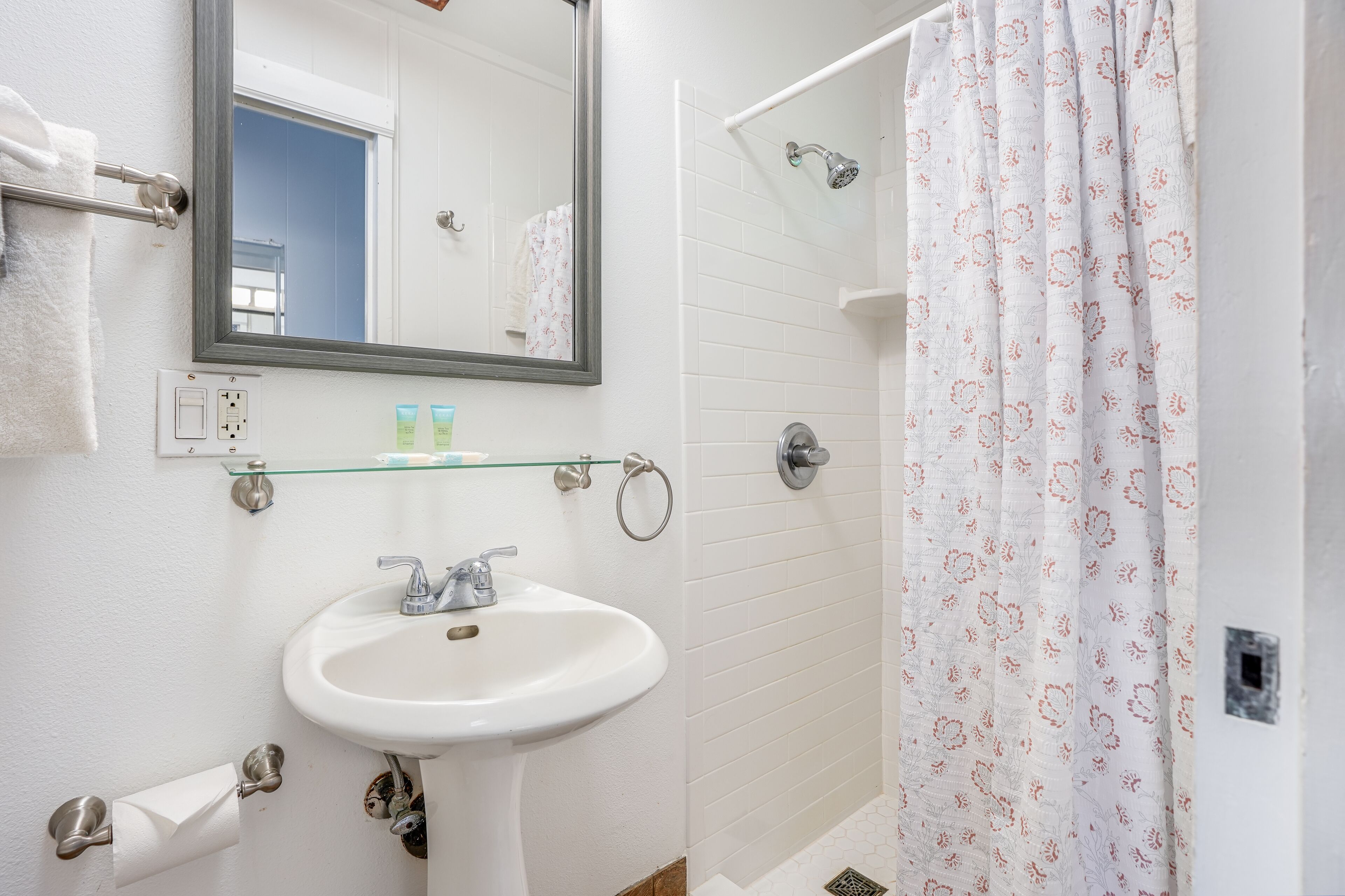 Une salle de bain avec un lavabo blanc, une étagère en verre au-dessus, un porte-serviettes et un rideau de douche à motifs floraux.