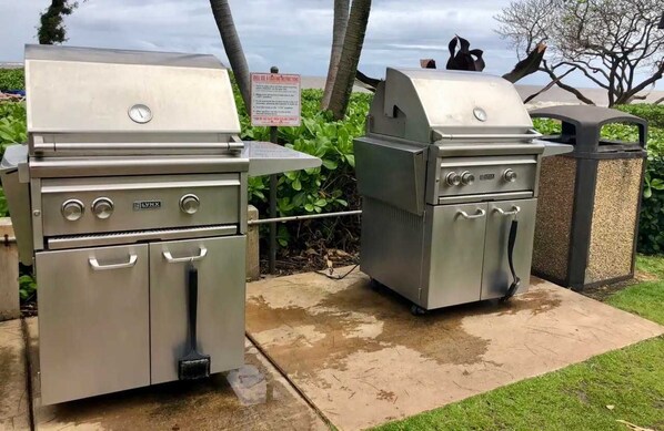 Two stainless steel barbecue grills with control knobs and a vent hood, placed on a concrete surface with a grassy area and trees in the background.